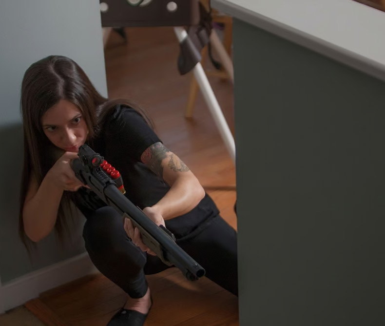 Woman crouching leaning looking down sights on her shotgun inside her home