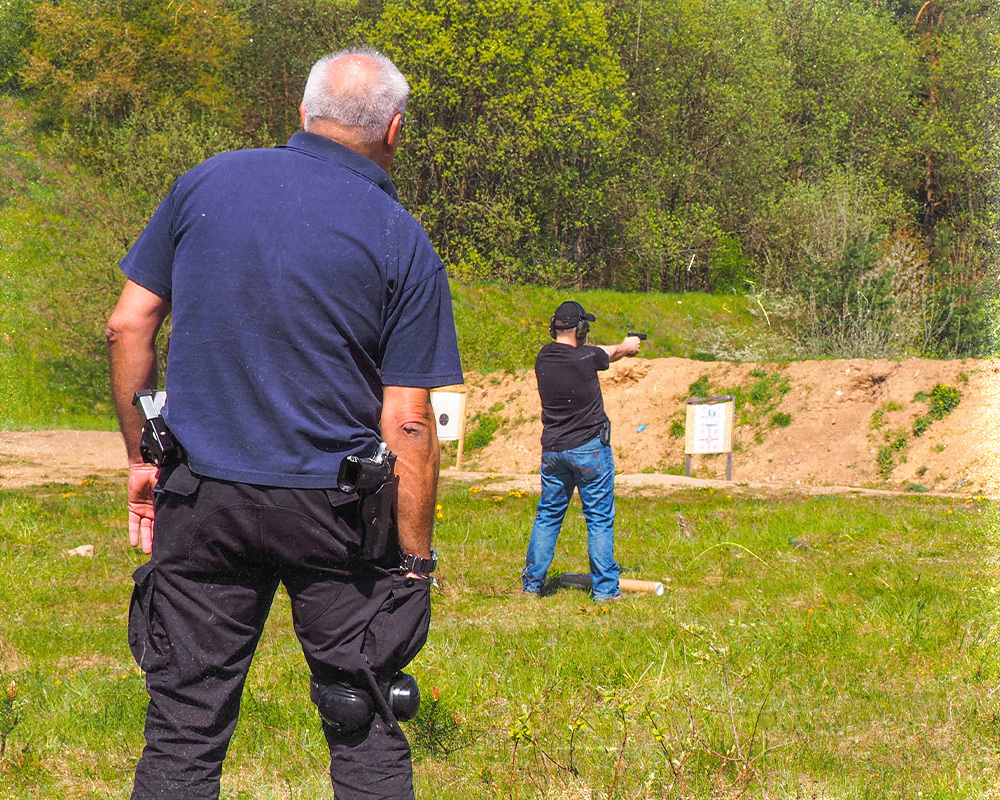 Elder man on a shooting range carrying both a OWB Holster and a Mag Holster, another man can be seen in the distance with a ready to shoot position taking shots at the targets in the distance.