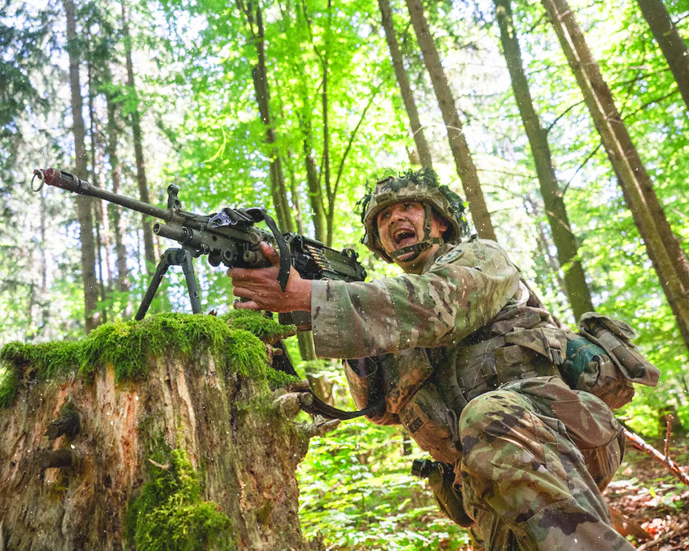 Soldier in camoflauge gear in a forest area with trees around him, holding a assault rifle.