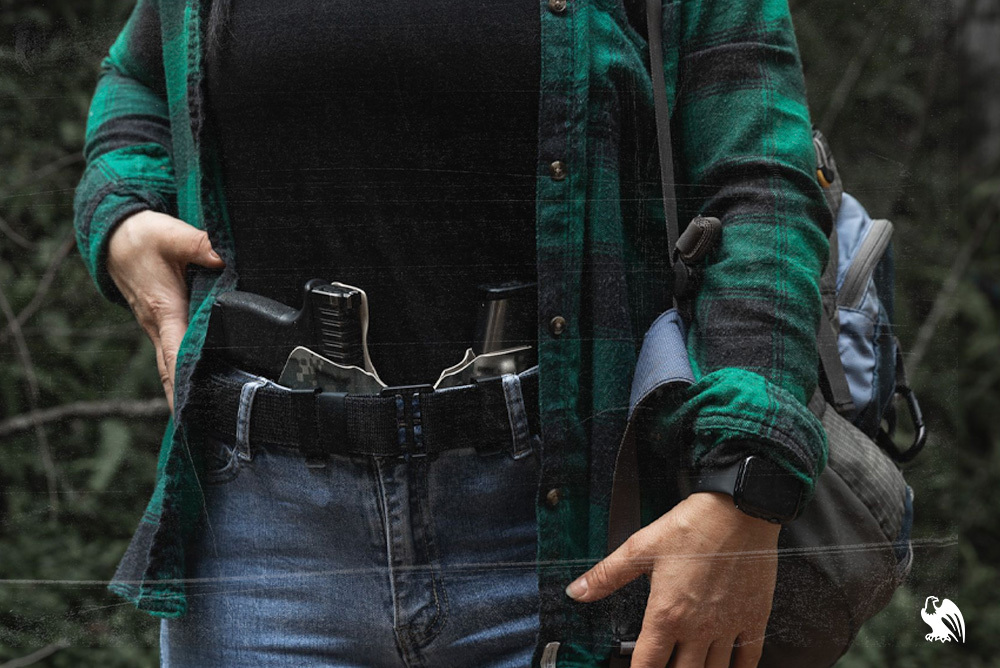 Woman wearing a flannel, black t-shirt, jeans, and a belt, carrying a backpack and a Vedder SideTuck IWB Kydex holster, showcasing practical concealed carry for everyday wear.
