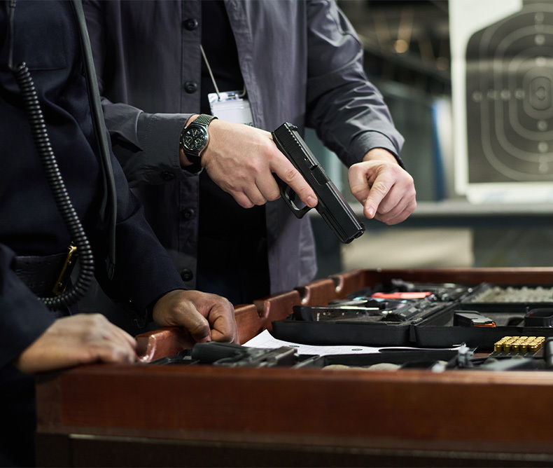 Two men inspecting guns layed on a table, in the background you can see a shooting target. 