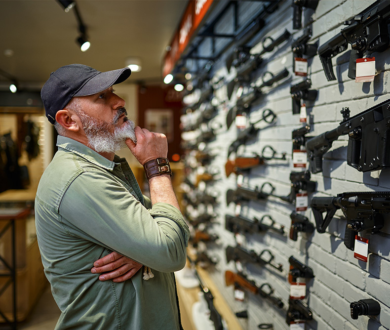 Man wearing a green shirt and a hat is checking a wall of guns, he is trying to decide which one to get.