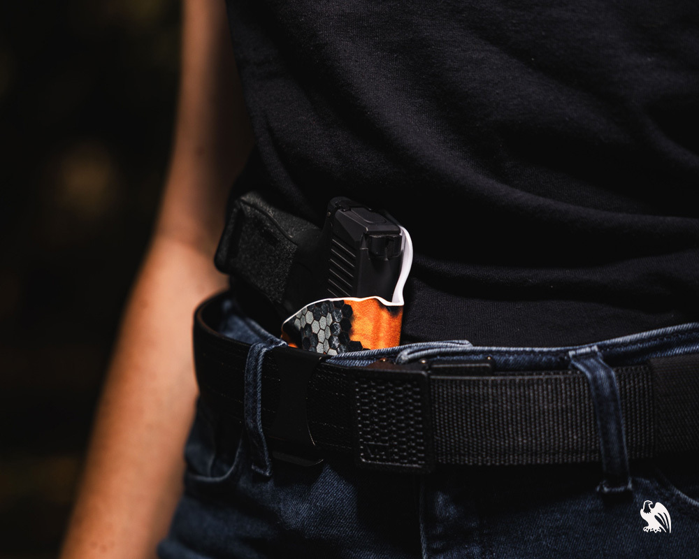 A close-up of an FN Reflex pistol holstered inside the waistband in a custom orange-patterned Vedder Kydex holster, worn with a reinforced gun belt.