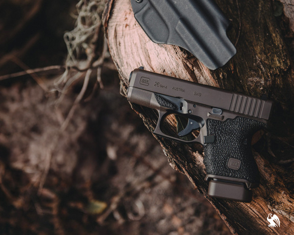 Glock 26 Gen4 9mm pistol with textured grip resting on a log beside a black Kydex holster in an outdoor woodland setting.