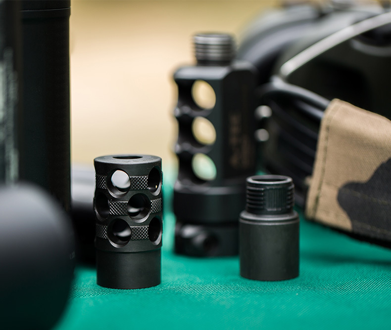 Close-up of various black muzzle devices, including compensators and thread adapters, displayed on a green surface, used to reduce recoil and enhance firearm performance.