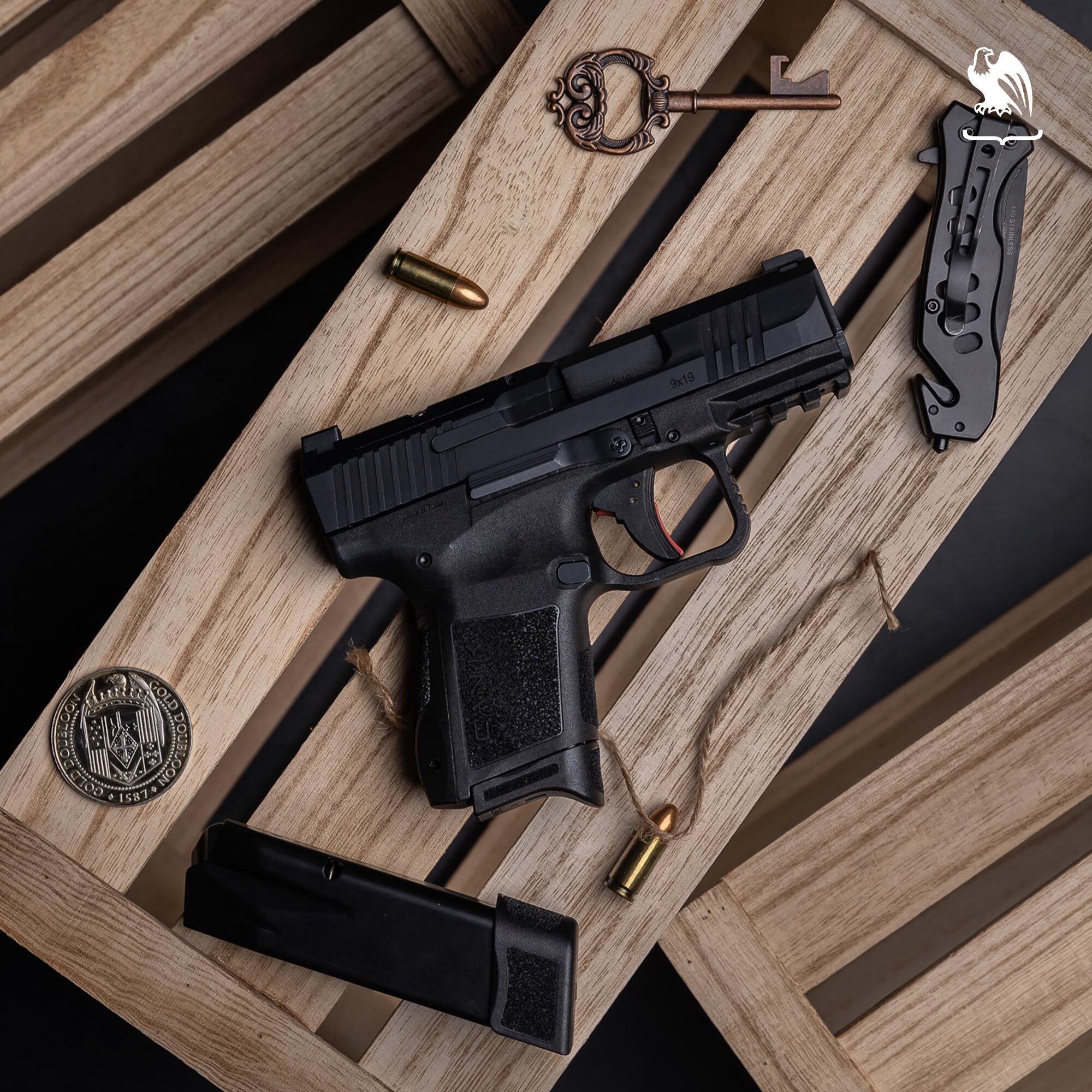 Canik MC9 9x19 compact pistol displayed on rustic wooden crates alongside a 9mm extended magazine, loose 9mm rounds, a folding tactical knife, an ornate antique key, and a challenge coin &mdash; overhead flat lay product photography.