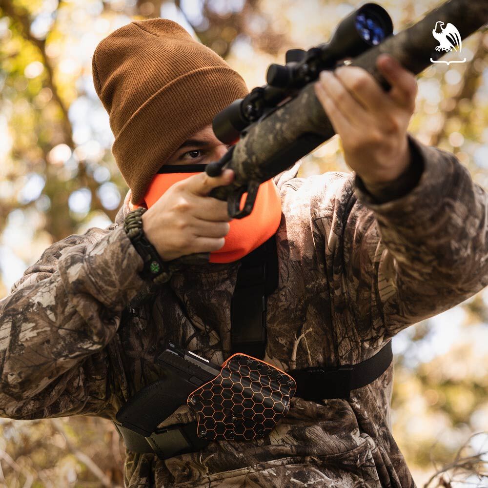 Hunter wearing camouflage gear and a beanie, aiming a rifle while carrying a handgun in a Evertrek Vedder Holsters chest holster.