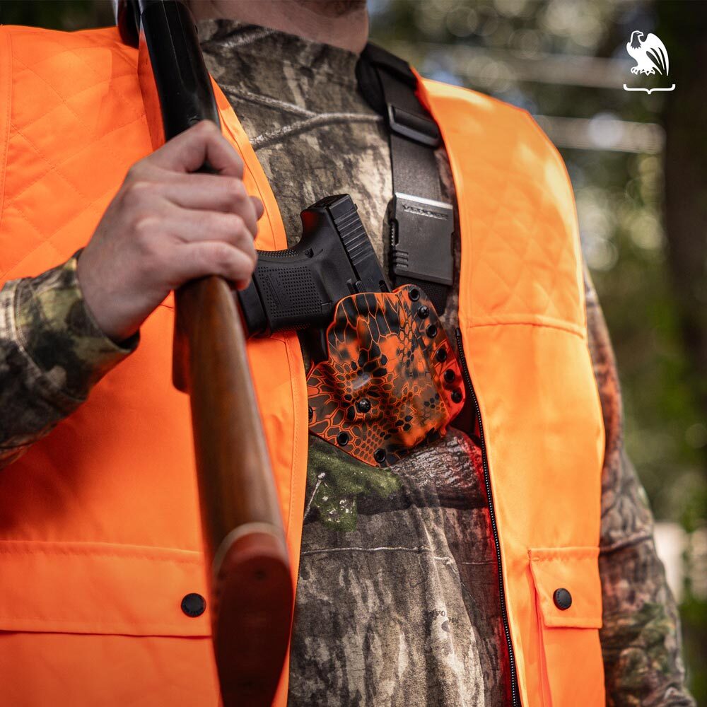 Hunter wearing blaze orange vest and camouflage clothing, carrying a shotgun with a handgun secured in a Evertrek Vedder Holsters chest holster.