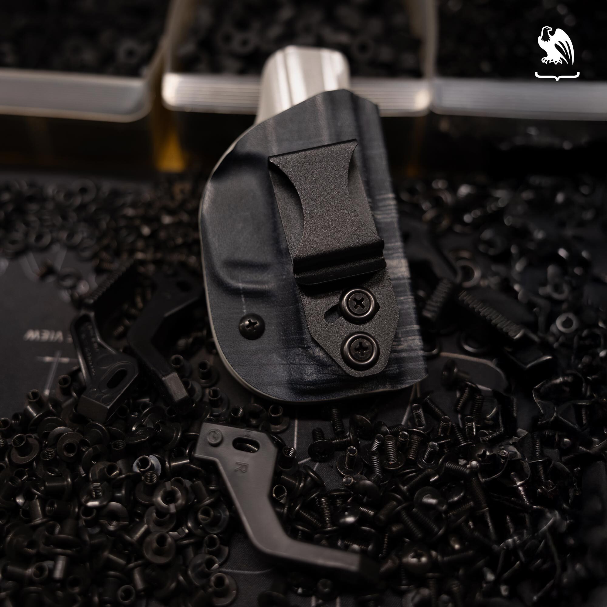 Custom Kydex IWB holster with sweat guard and adjustable clip surrounded by loose holster hardware including screws, nuts, and eyelets on a dark workshop surface &mdash; handmade Kydex holster customization and hardware components.
