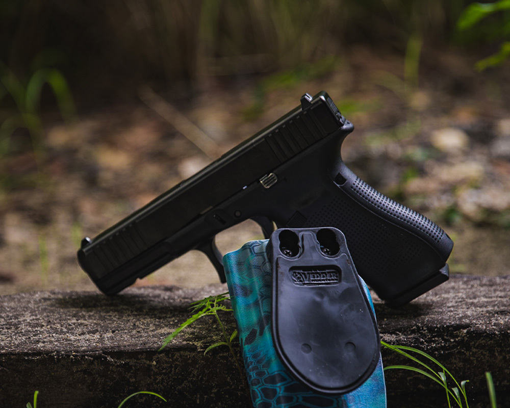 Black handgun resting on a log beside a custom blue Kydex Vedder Holster, photographed outdoors with a natural background