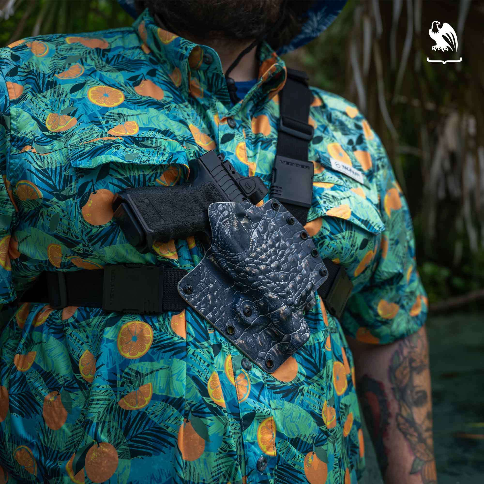 Chest Holsters Ideal for Hiking A man in a green, blue, and orange Hawaiian shirt carries a pistol in a Vedder Holsters EverTrek Chest Holster with a gator skin Kydex pattern.