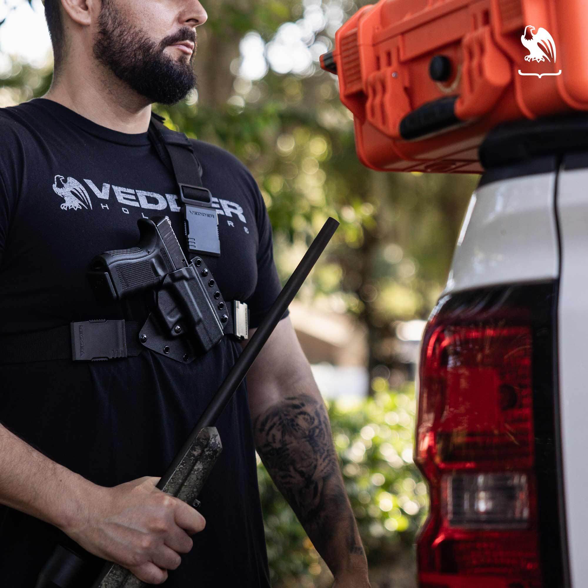 Man wearing a Vedder Holsters chest holster with a handgun secured, holding a hunting rifle beside a truck.