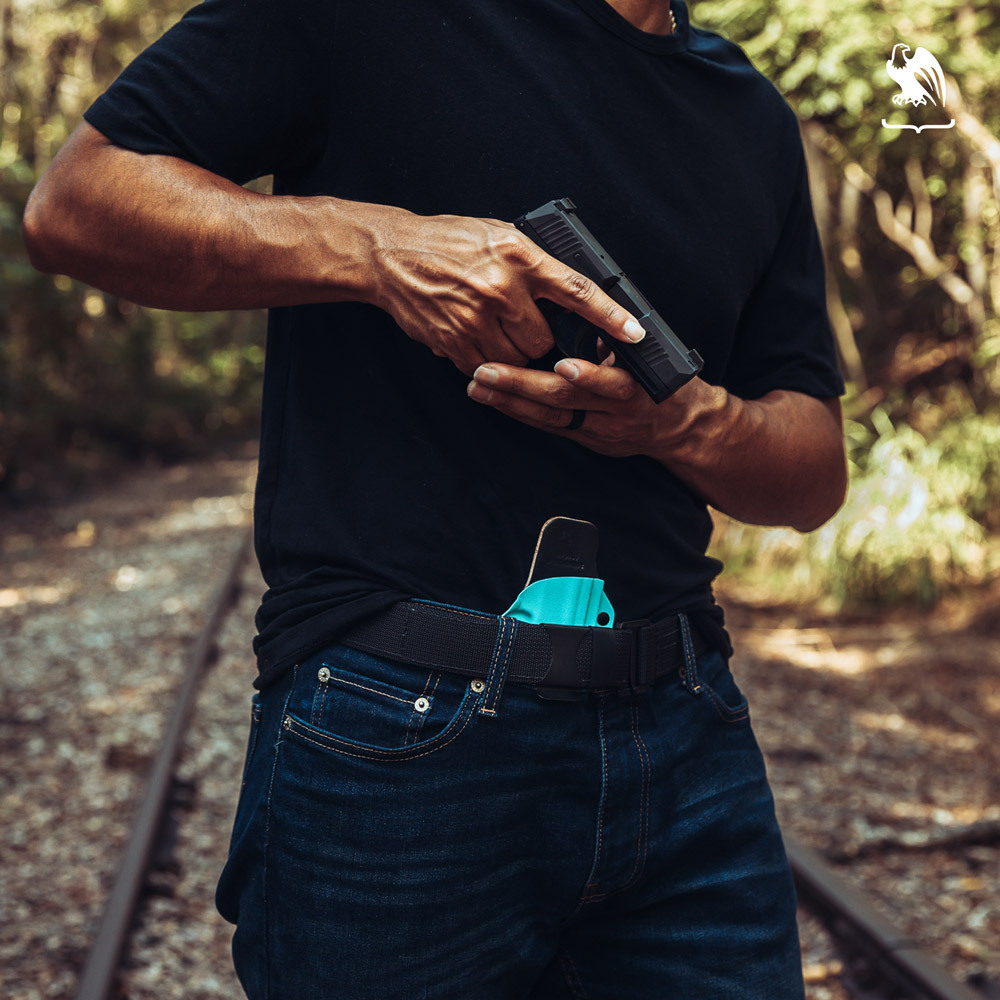 Man wearing jeans and a black shirt drawing a handgun from a turquoise Kydex IWB concealed carry holster secured with a gun belt.