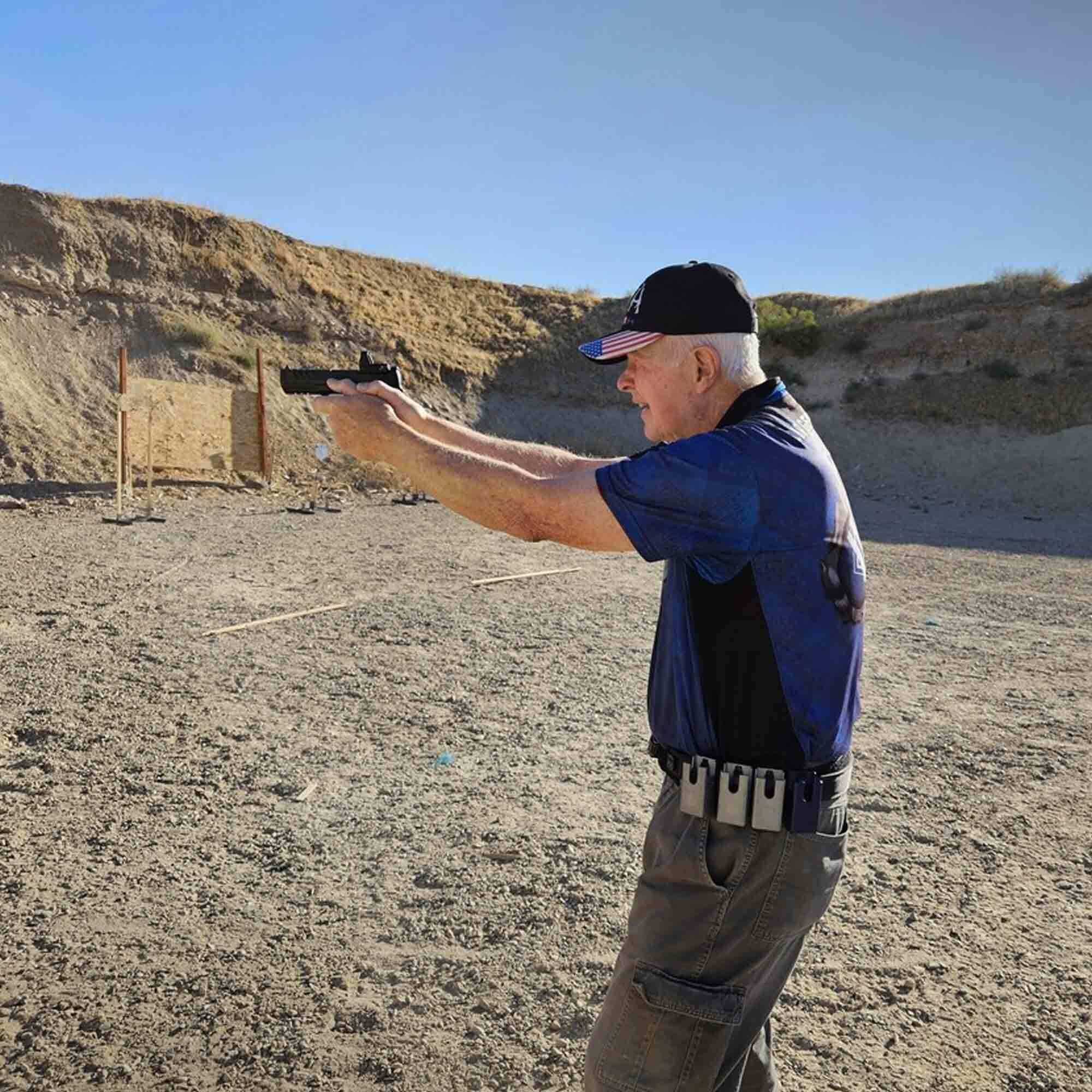 Navy veteran Rick Junge practices shooting at an outdoor range, aiming his pistol with proper stance and technique. He wears tactical gear with extra magazines on his belt, showcasing firearm training and marksmanship skills.