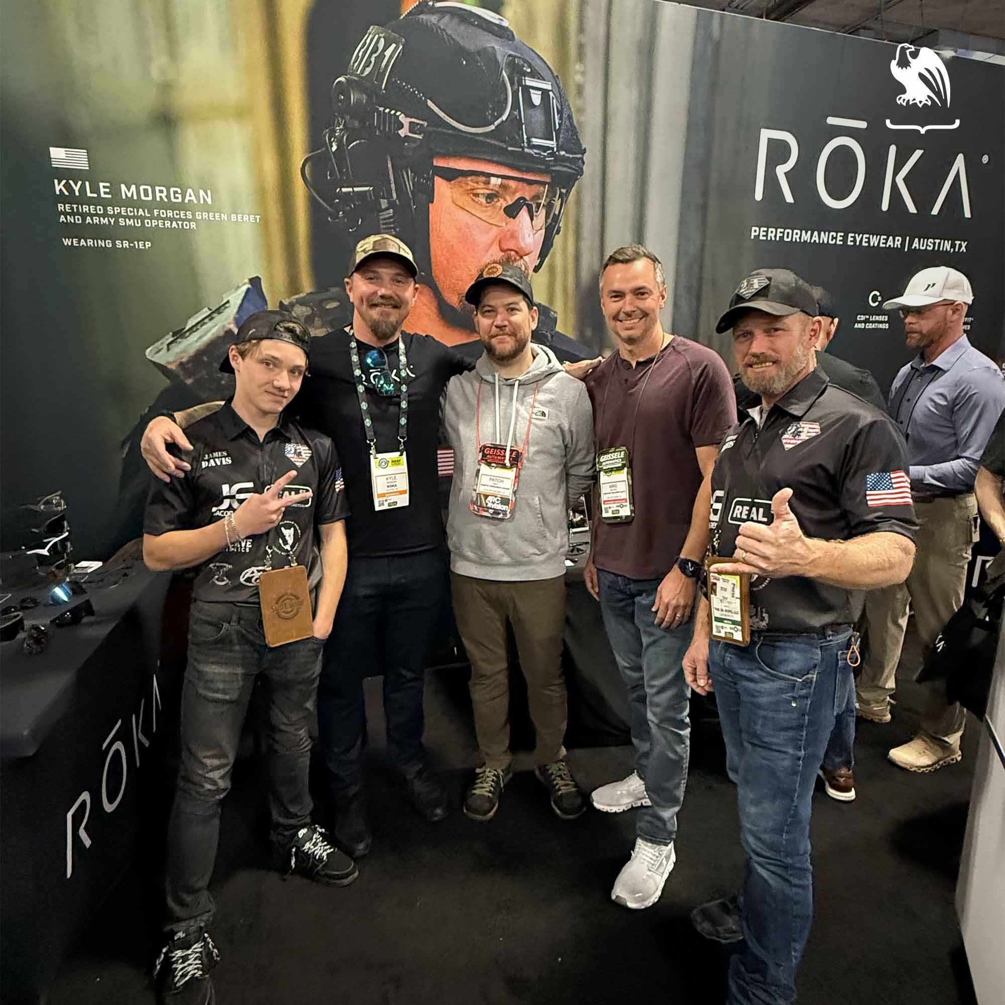 Group of firearms and tactical industry professionals networking at the ROKA Performance Eyewear booth during SHOT Show, with ballistic eyewear branding and military-themed display in the background.