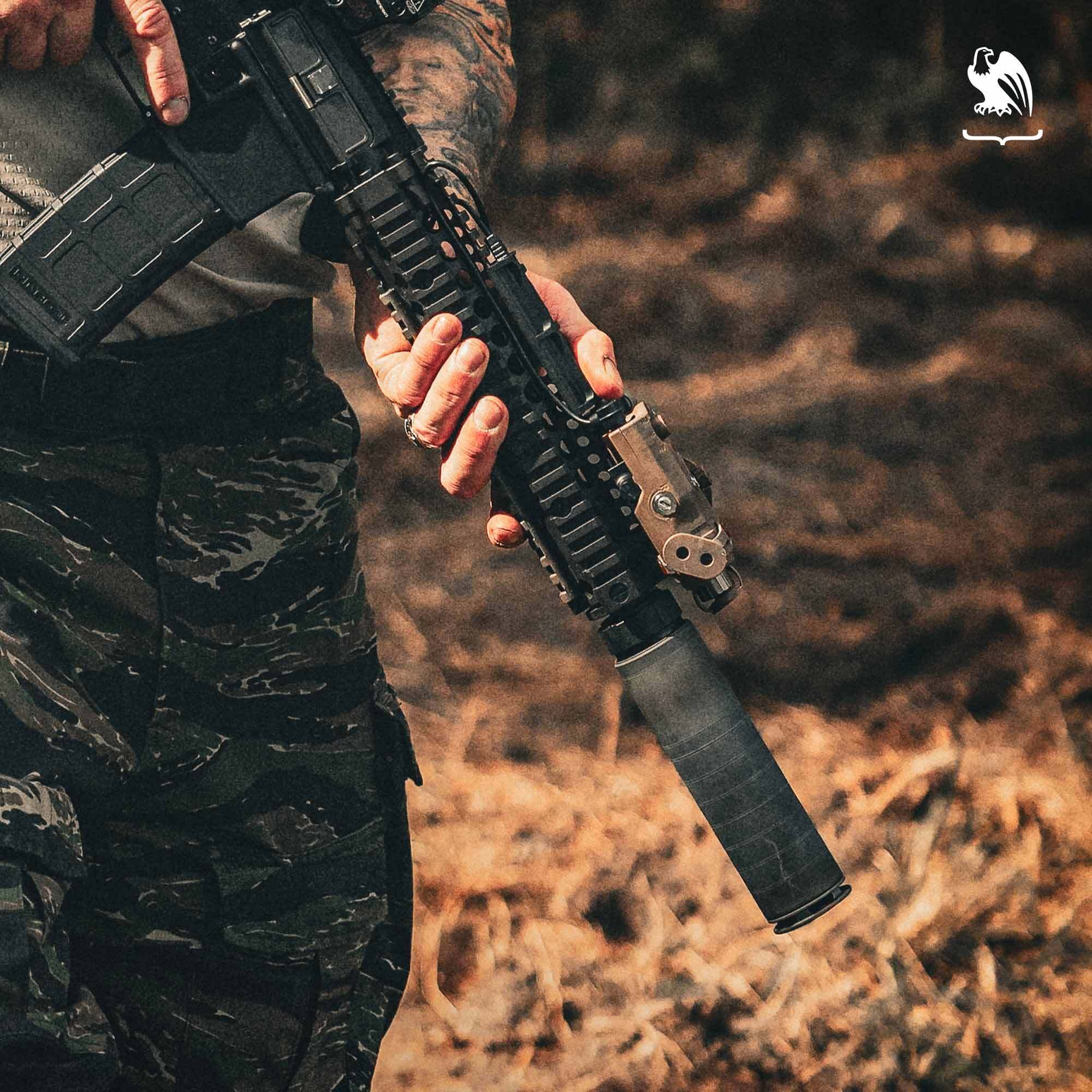 Person holding a suppressed AR-style rifle outdoors, showing a mounted suppressor during a shooting or training scenario