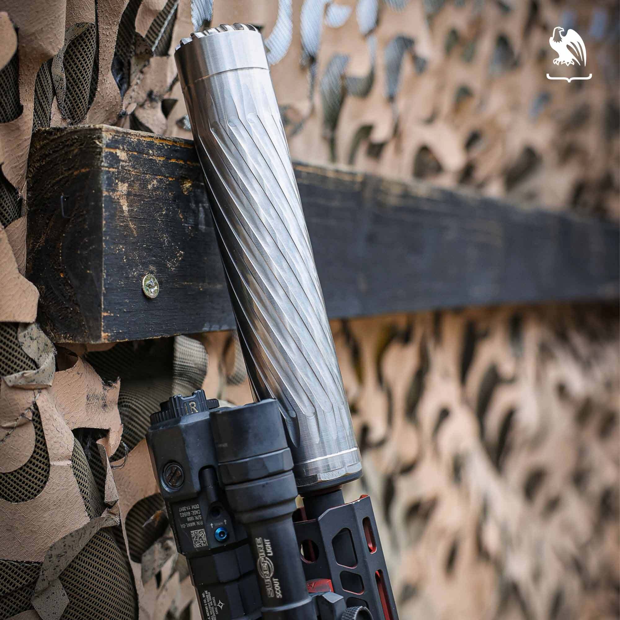 Close-up of a stainless steel rifle suppressor mounted on a tactical firearm against a camouflage background.