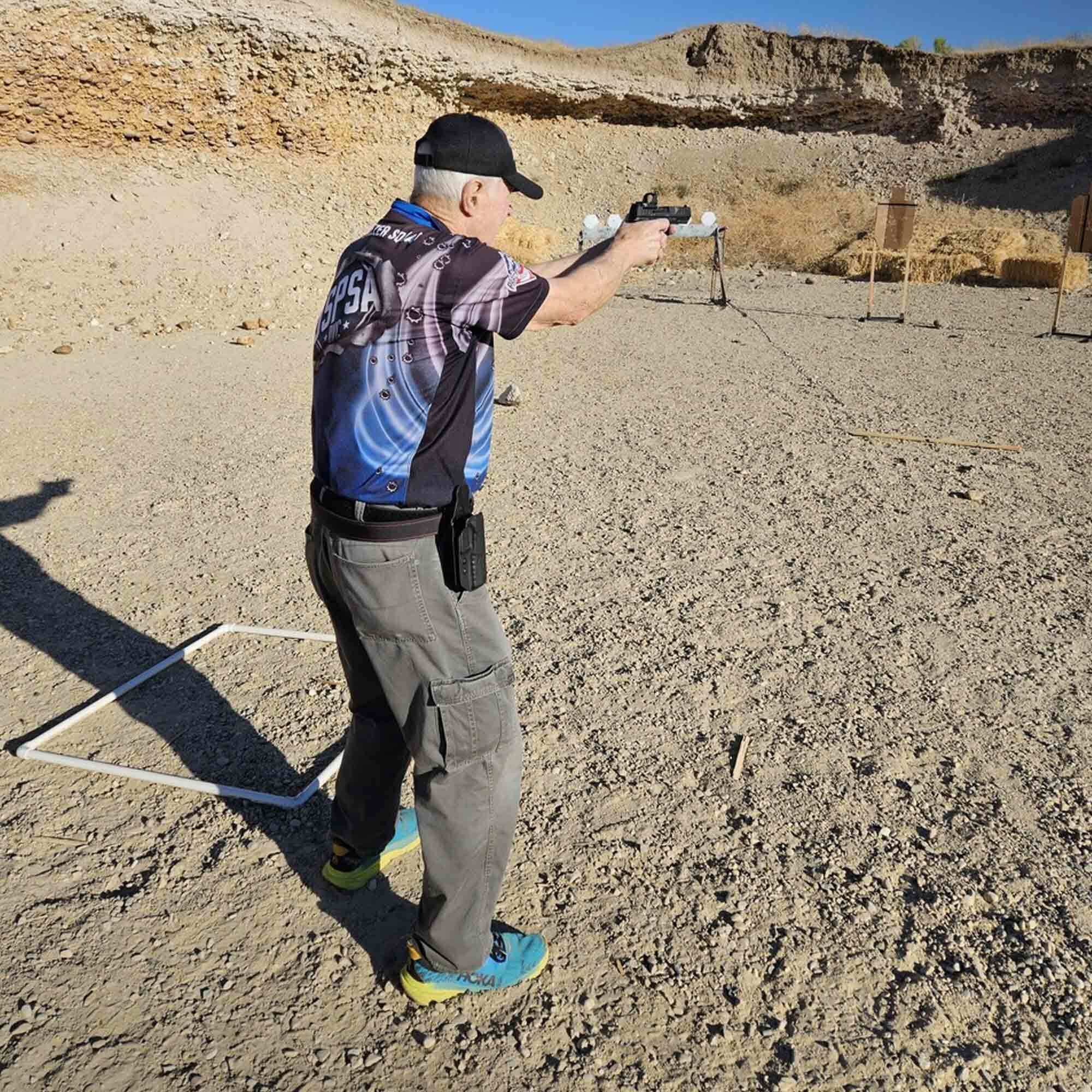 Veteran Rick Junge competes in a shooting match at an outdoor range, aiming his pistol at targets while wearing a USPSA jersey, tactical pants, and holster gear.