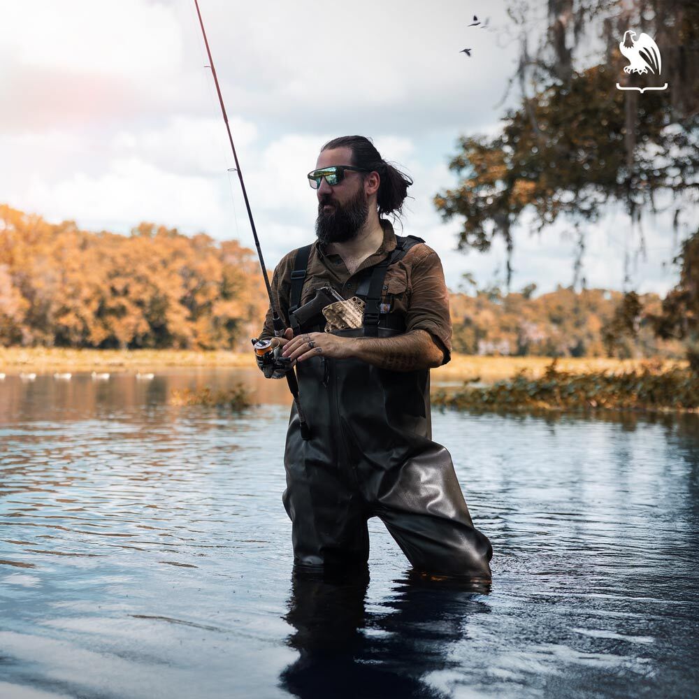 Man fishing in knee-deep water wearing waders and sunglasses, carrying a handgun in a Vedder Holsters Evertrek chest holster.